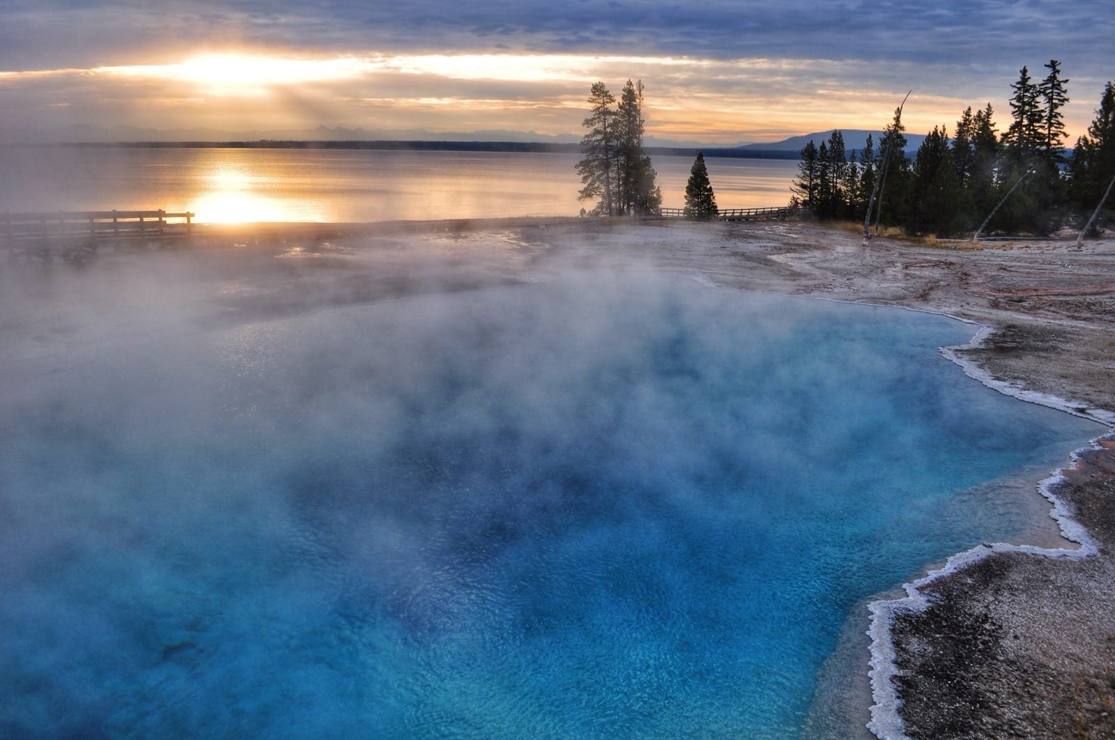 Color photo of the sun rising over Yellowstone Lake with a bright blue geyser in the foreground.